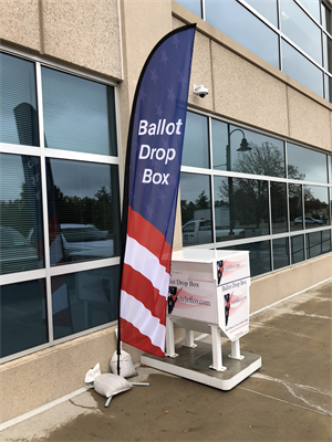 Ballot box on a sidewalk next to windows with a red white and blue ballot drop box flag next to it.